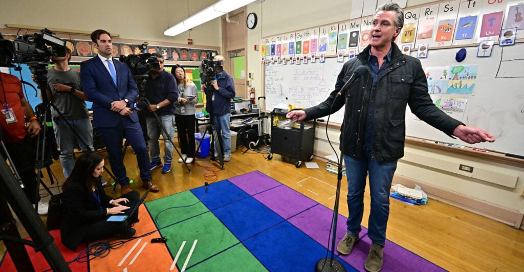 Gavin Newsom addresses the press while in an elementary school classroom, standing on a rainbow -colored rug.
