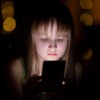 Young girl with serious expression, her face lit by a smartphone against dark background.