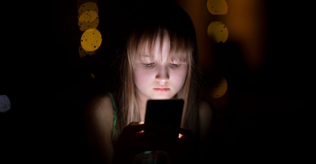 Young girl with serious expression, her face lit by a smartphone against dark background.