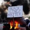 Someone holds up sign reading "Black Lives Matter" in front of burning police car.