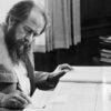 Black-and-white photo of Russian author Aleksandr Solzhenitsyn taking notes at a desk with a large manuscript in front of him.