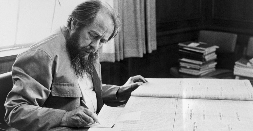Black-and-white photo of Russian author Aleksandr Solzhenitsyn taking notes at a desk with a large manuscript in front of him.