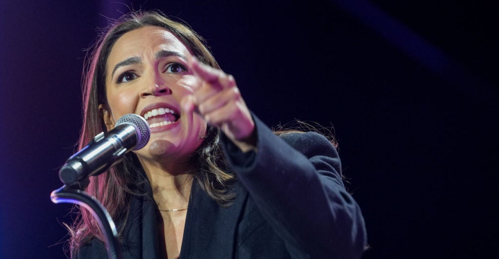 Rep. Alexandria Ocasio-Cortez, in a blue suit, points while delivering a speech against a dark background.