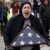 Woman in black winter coat with hoodie screams at ICE agents while holding folded American flag.
