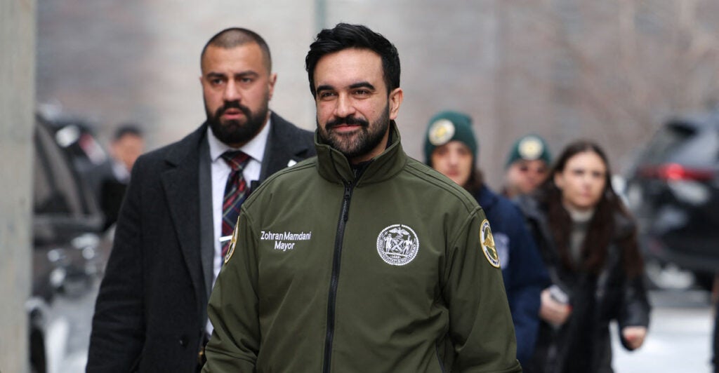 Zohran Mamdani walks outside while wearing a green winter jacket.