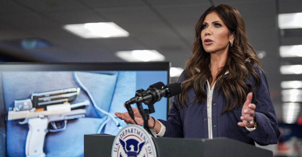 DHS Secretary Kristi Noem in a blue jacket speaks behind a podium next to a photo of a semi-automatic handgun.
