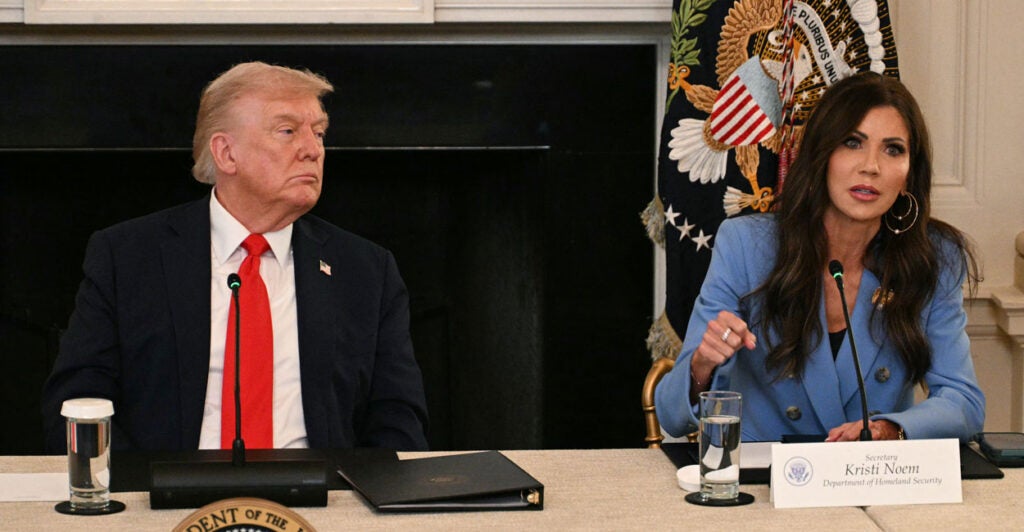 Donald Trump listens to DHS Secretary Kristi Noem as they sit at a table.