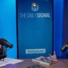 Jack Fowler and Tyler O'Neil talk at a studio table against a blue backdrop with screen reading "The Daily Signal."
