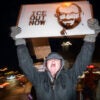Woman in gray winter clothing screams as she holds up sign with image of Alex Pretti with caption ICE OUT NOW."