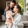 Parents share a joyful moment in their living room while holding their baby girl. The space is bright and welcoming with soft furniture and green plants around them.