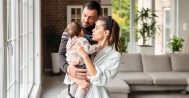 Parents share a joyful moment in their living room while holding their baby girl. The space is bright and welcoming with soft furniture and green plants around them.