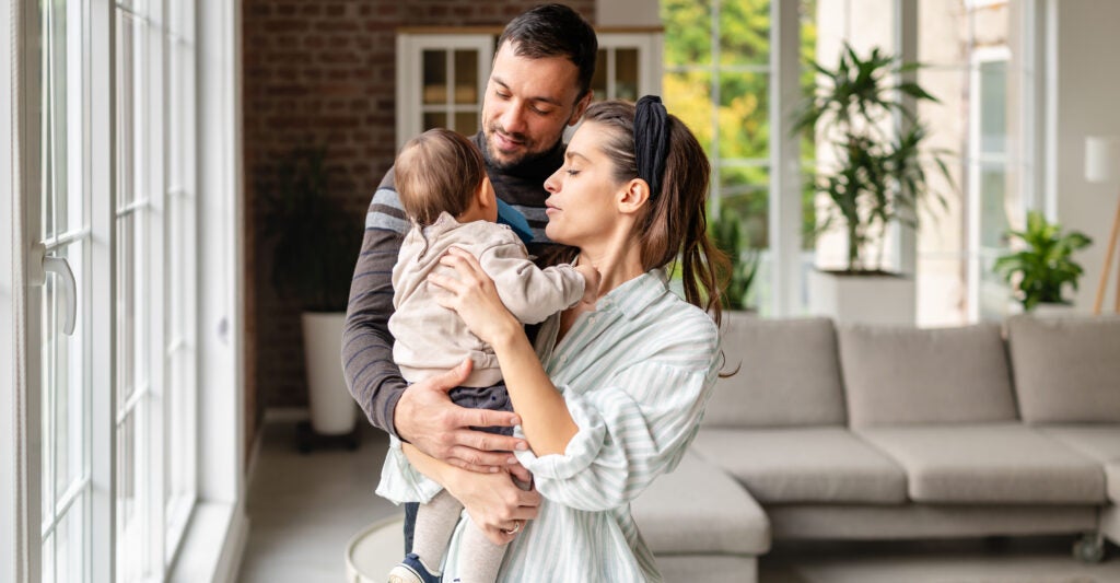 Parents share a joyful moment in their living room while holding their baby girl. The space is bright and welcoming with soft furniture and green plants around them.