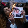 An anti-Iranian regime protester lights a cigarette with a lit paper depicting Ali Khamenei, Supreme Leader of Iran during a gathering.