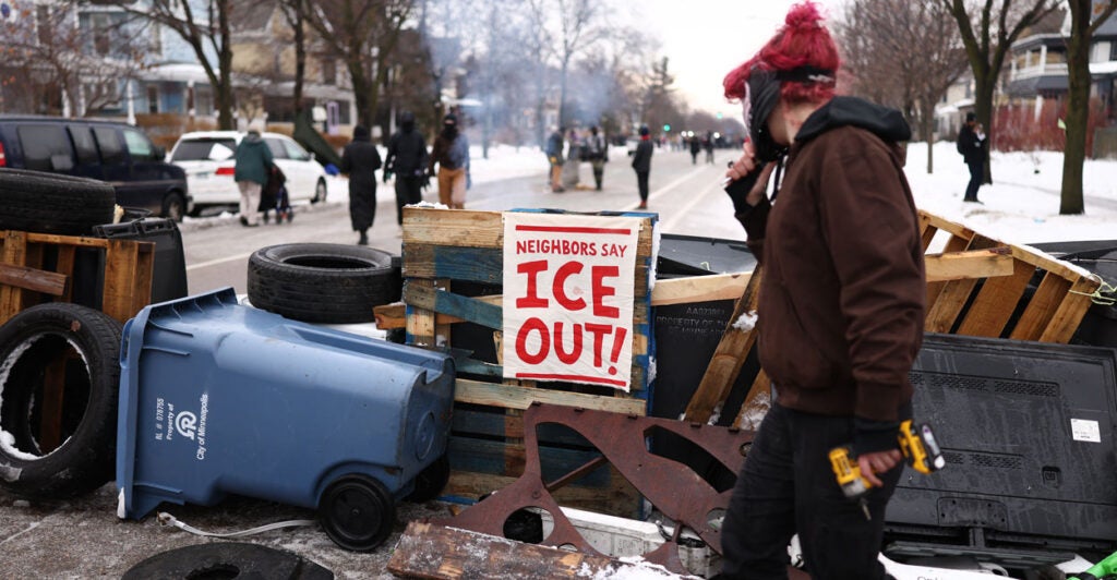 Makeshift barricade set up by anti-ICE protesters blocks Minneapolis street. On barricade is sign reading "Neighbors say ICE Out." Protester is crossing on right of image.