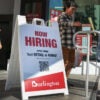 Older woman, young man and woman walk around Burlington "Now Hiring" sign outside a store.