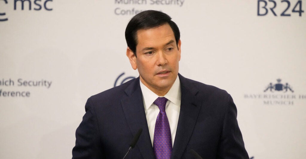 Marco Rubio in a dark suit and purple tie speaks against a white background.