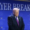 President Trump bows his head in prayer in front of blue "Prayer Breakfast" background.