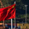 CHONGQING, CHINA - AUGUST 12: The national flag of China waves in the wind at a container terminal on the Yangtze River, with dockside cranes and stacked shipping containers visible in the background, on August 12, 2025 in Chongqing, China. (Photo by Cheng Xin/Getty Images)