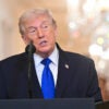 President Trump in dark suit and blue tie speaks at a podium in East Room of the White House.