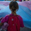 Little child with red backpack and teddy bear stands looking at a trans flag.