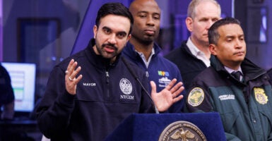 Zohran Mamdani in blue jacket stands at podium with arms outstretches and officials look on.