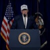 President Donald Trump behind a podium, in blue jacket, white shirt and "USA" cap, speaks.