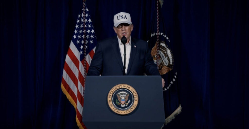 President Donald Trump behind a podium, in blue jacket, white shirt and "USA" cap, speaks.