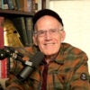 A smiling shot of Victor Davis Hanson seated at a microphone in a rust-colored flannel shirt.