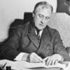 Black-and-white photo of President Franklin D. Roosevelt at desk signing legislation.