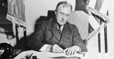 Black-and-white photo of President Franklin D. Roosevelt at desk signing legislation.