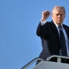 President Donald Trump raises his fist at the top of the stairs to Air Force One against a perfect blue sky.