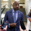 Sen. Raphael Warnock, in blue suit talks to reporters inside the U.S. Capitol.