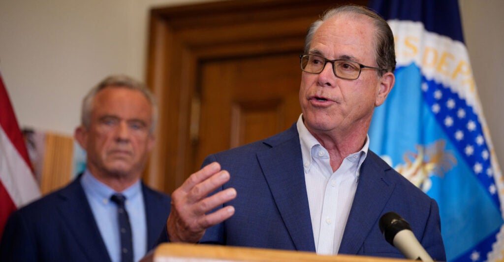 Indiana Gov. Mike Braun gestures while speaking at a podium, HHS Secretary Robert F. Kennedy Jr. behind him.