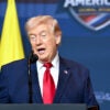 President Trump gestures with his right hand while speaking at a podium in front of a "Shield of America" sign.