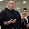 John Fetterman in black hoodie holds cellphone while talking to reporters.