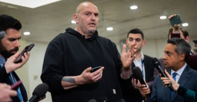 John Fetterman in black hoodie holds cellphone while talking to reporters.