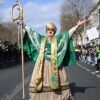 An actor depicting St. Patrick spreads his arms wide in the middle of a Dublin street as parade spectators look on.