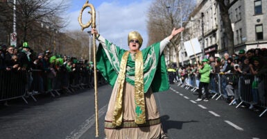 An actor depicting St. Patrick spreads his arms wide in the middle of a Dublin street as parade spectators look on.