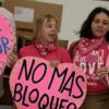 MIAMI, FLORIDA - MARCH 20: (L-R) Medea Benjamin, Jazmin Rumbaut,and Michelle Ellner from CODEPINK hold signs as they check in boxes of humanitarian aid at Invicta Air Charter in the Miami International Airport on March 20, 2026 in Miami, Florida.