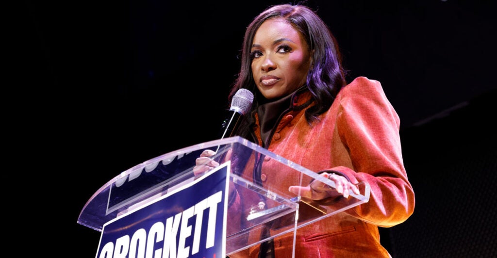 Rep. Jasmine Crockett in red suit looks out sternly from behind podium bearing her name.