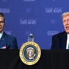 Secretary of War Pete Hegseth watches President Donald Trump speak in front of a blue backdrop with the words in white "Make America Safe Again."