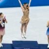 Alysa Liu leaping in the air on the Olympic podium as two other skaters applaud.