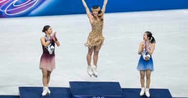 Alysa Liu leaping in the air on the Olympic podium as two other skaters applaud.