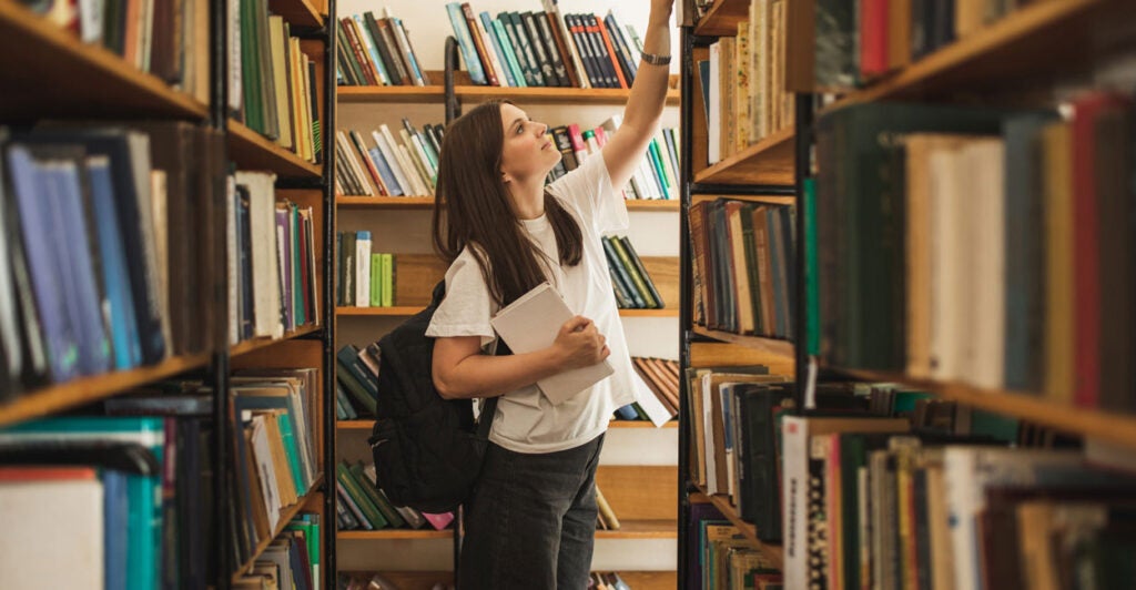 Female student reaching up for a book in a school library stack.