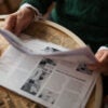 Unseen older person reading a daily newspaper while sitting at round table with cup of coffee