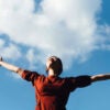Woman outside in red shirt with her arms outstretched.