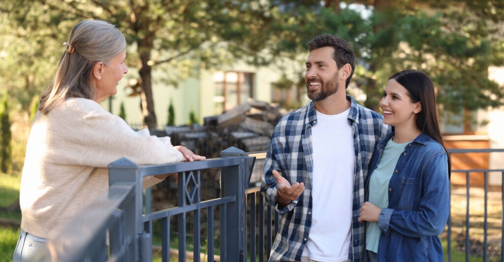 Friendly relationship with neighbors. Happy young couple talking to senior woman near fence outdoors.