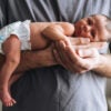Male in gray T-shirt holds a newborn, whose legs dangleoff his arm.