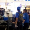 TSA agents assisted by ICE agents screen passengers at an airport.