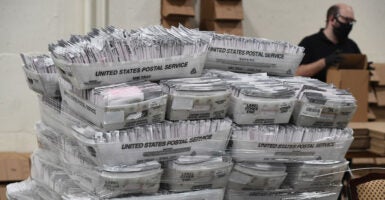 Large stack of bins filled with mail-in ballots, with postal worker in background.